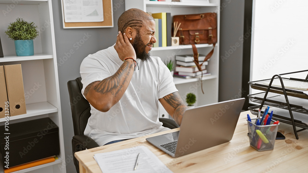 African american man with beard feeling neck pain at office workstation with laptop and decorative plant in background - obrazy, fototapety, plakaty 