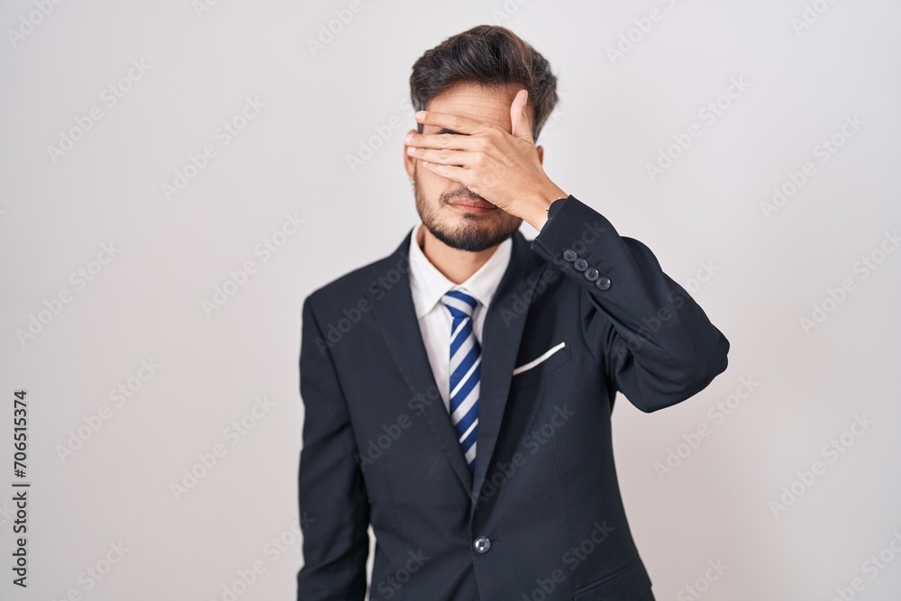Young hispanic man with tattoos wearing business suit and tie covering eyes with hand, looking serious and sad. sightless, hiding and rejection concept Young hispanic man with tattoos wearing business suit and tie covering eyes with hand, looking serious and sad. sightless, hiding and rejection concept