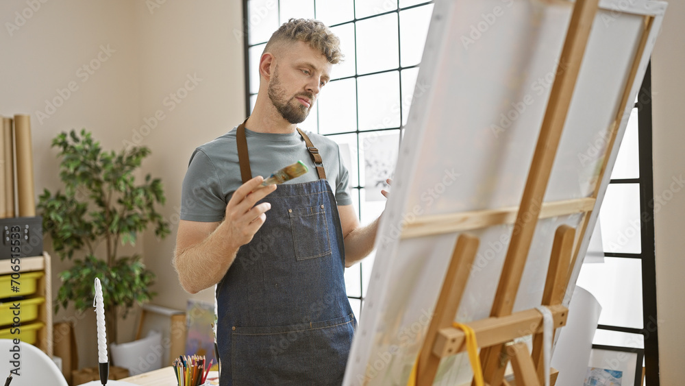 Fototapeta premium Young caucasian man with a beard and blue eyes, wearing a denim apron, thoughtfully painting on an easel in a brightly lit studio