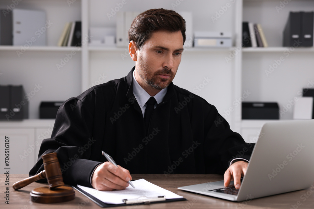 Judge with gavel and laptop writing in papers at wooden table indoors
