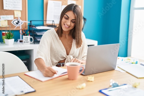 Young beautiful hispanic woman business worker using laptop writing on notebook at office