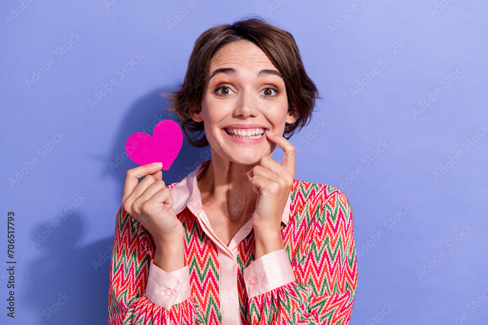 Photo portrait of lovely young lady bite nail hold pink heart postcard wear trendy print garment isolated on violet color background Photo portrait of lovely young lady bite nail hold pink heart postcard wear trendy print garment isolated on violet color background