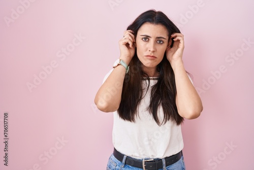 Young brunette woman standing over pink background covering ears with fingers with annoyed expression for the noise of loud music. deaf concept.