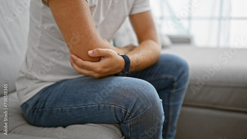 A young hispanic man experiencing elbow pain while sitting on a couch indoors.