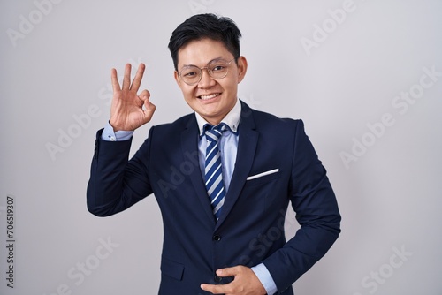 Young asian man wearing business suit and tie smiling positive doing ok sign with hand and fingers. successful expression.
