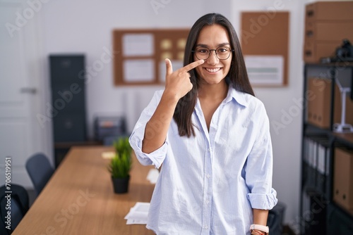 Young hispanic woman at the office pointing with hand finger to face and nose, smiling cheerful. beauty concept photo