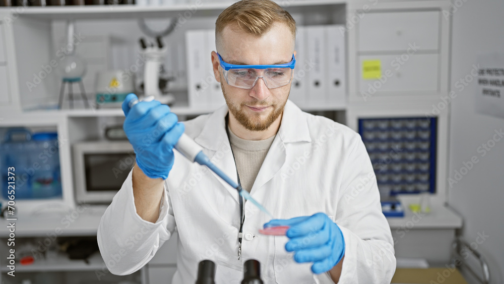 A focused young man in labcoat performs an experiment in a laboratory, pipetting a blue substance into a petri dish. A focused young man in labcoat performs an experiment in a laboratory, pipetting a blue substance into a petri dish.