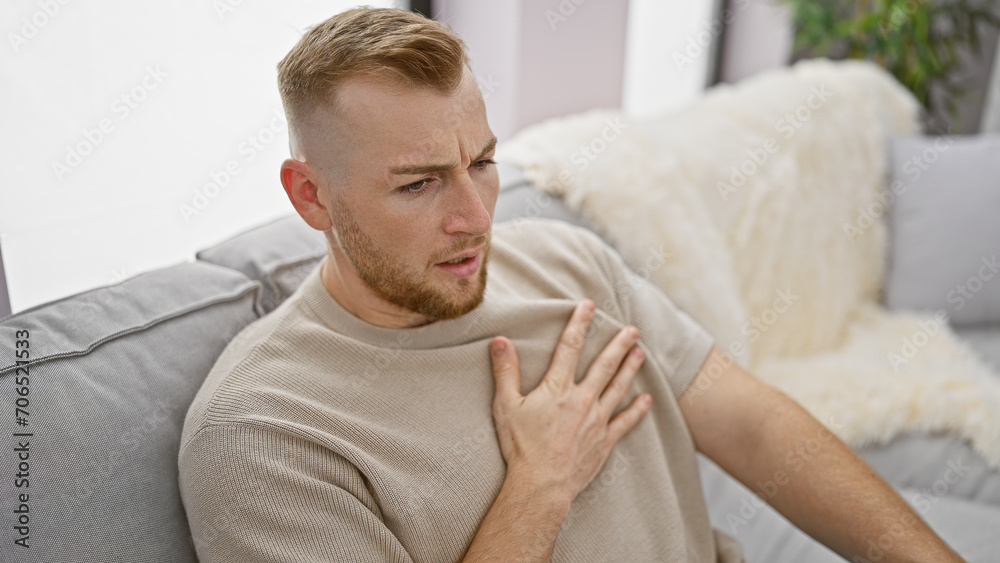 Caucasian bearded man experiencing chest pain while sitting on a couch indoors. Caucasian bearded man experiencing chest pain while sitting on a couch indoors.