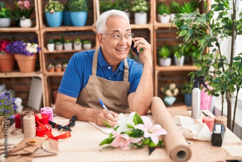 Middle age grey-haired man florist talking on smartphone writing on notebook at florist