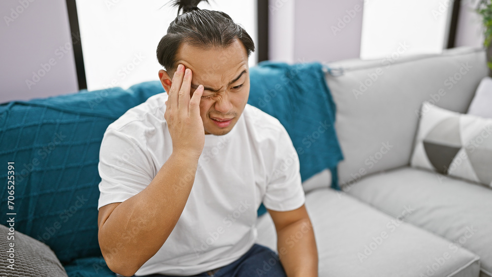 Stressed young chinese man, frustrated and alone, sits on sofa nursing a headache at home amidst sadness and personal conflict Stressed young chinese man, frustrated and alone, sits on sofa nursing a headache at home amidst sadness and personal conflict