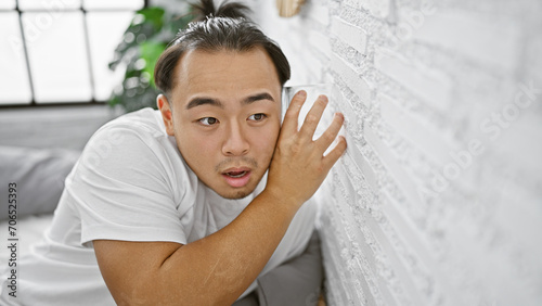 Nosy young chinese man sneakily listening through bedroom wall with glass, overcome by curiosity and eavesdropping in a secret indoor spy mission in his apartment. photo