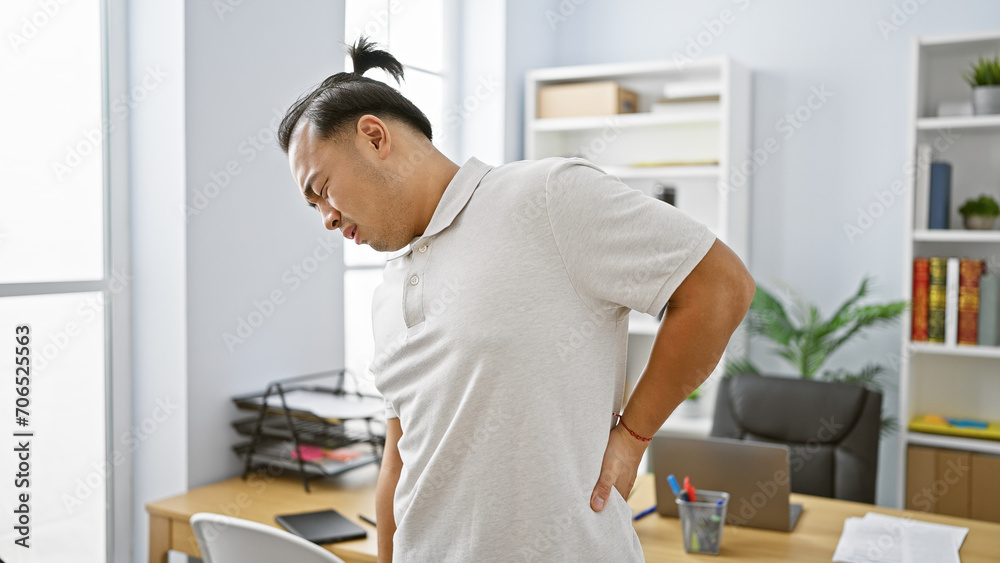 Worried young chinese business worker suffering severe backache, while working tirelessly indoors at the office, showing signs of spinal injury