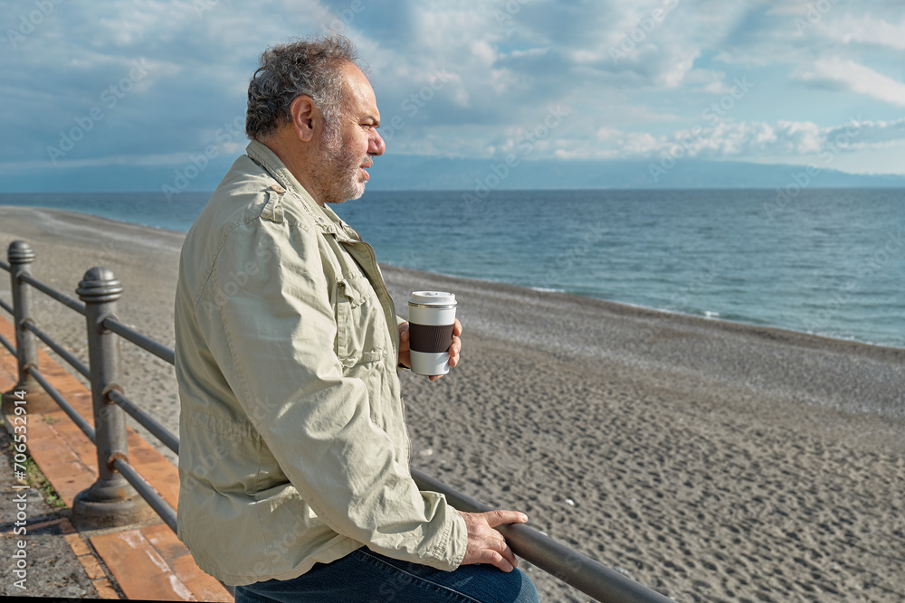 Pensive middle-aged bearded man drinking coffee on seaside in sunny winter or spring day. Pensive middle-aged bearded man drinking coffee on seaside in sunny winter or spring day.