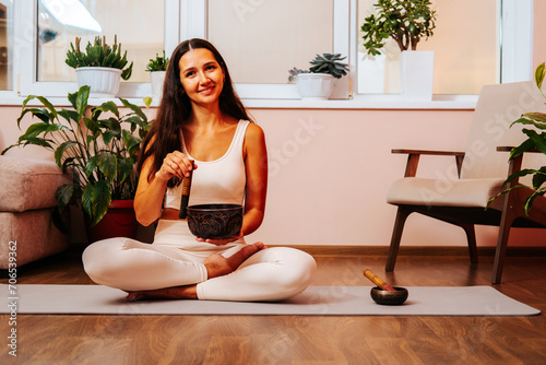 Happy beautiful young woman is sitting in lotus position and palyying singing bowl on yoga mat at yoga class. Soundhealing concept. © dvulikaia
