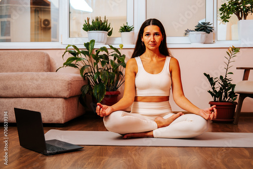 Beautiful young brunette woman is sitting in lotus position on yoga mat at home in living room.