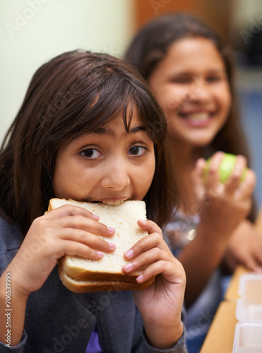 Hungry girl, portrait and student eating sandwich in classroom at school for meal, break or snack time. Young kid or elementary child biting bread for lunch time, fiber or nutrition in class recess photo