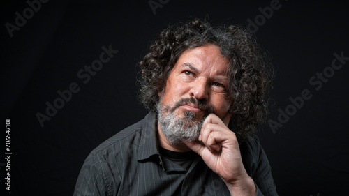 Man with white beard and black curly hair with thoughtful expression, resting his head on his hand, wearing black shirt against black background