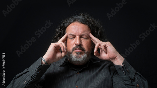 Man with white beard and black curly hair with concentrated expression, with his fingers on his temples in his hand, wearing black shirt against black background