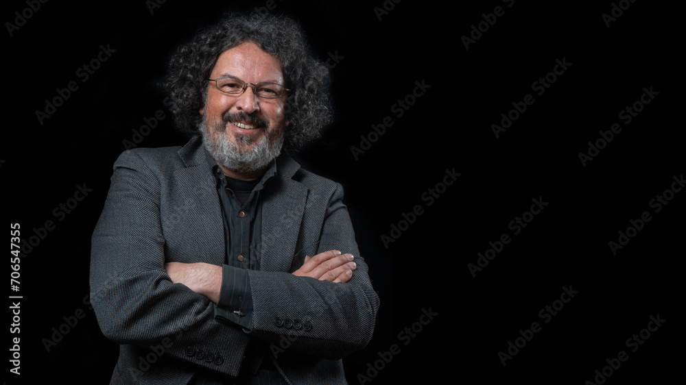 Man with white beard and black curly hair with happy expression, smiling towards camera, wearing black shirt against black background