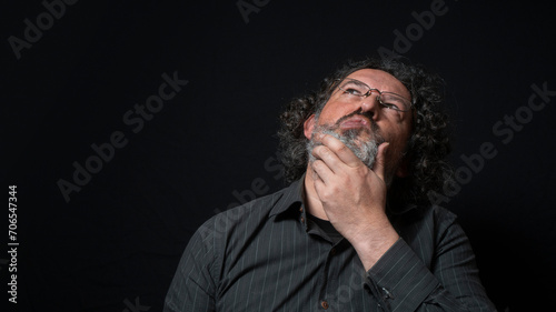 Man with white beard and black curly hair with expression of indecision and doubt, looking up, wearing black shirt against black background