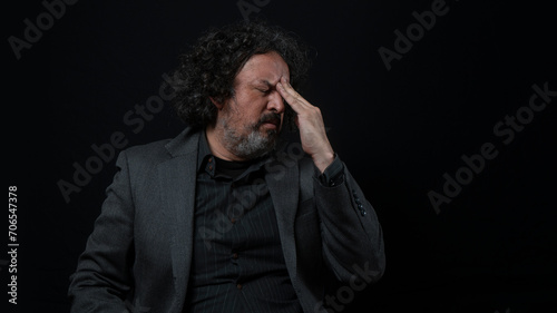 Man with white beard and black curly hair with pained expression, with his hand on his forehead, wearing black shirt against black background