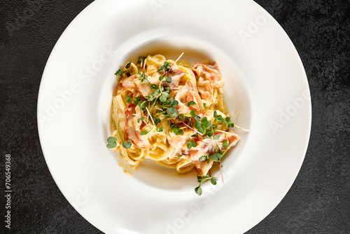Creamy carbonara pasta topped with crispy bacon and fresh microgreens on a white plate, against a dark backdrop