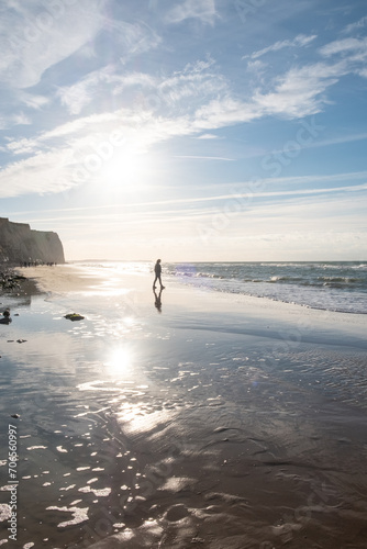 This photograph captures a solitary figure taking a peaceful walk along the shore, bathed in the ethereal glow of the coastal sunlight. The sun, hanging low in the sky, casts a brilliant reflection on