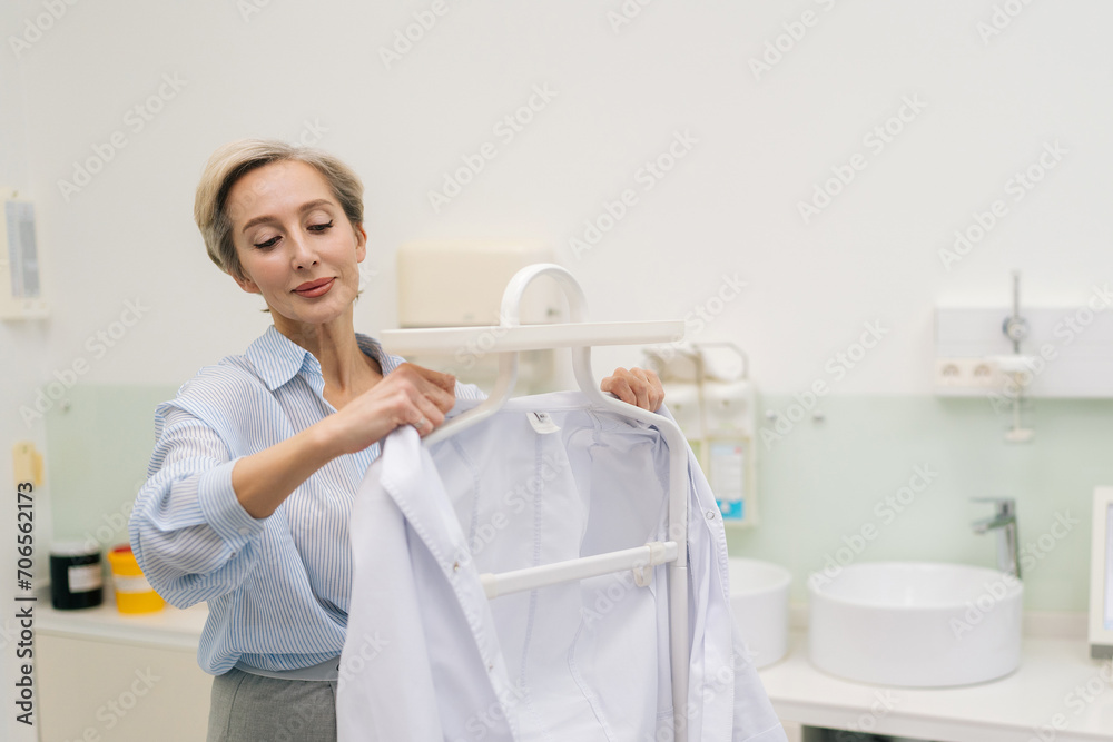 Front view of middle-aged female doctor walks into office, takes robe off coat hanger and puts it. Professional practitioner getting ready to work in clinic with modern interior. Concept of healthcare Front view of middle-aged female doctor walks into office, takes robe off coat hanger and puts it. Professional practitioner getting ready to work in clinic with modern interior. Concept of healthcare