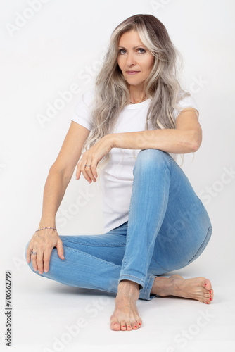 A beautiful elderly woman with gray long hair in jeans poses while sitting on a white background.