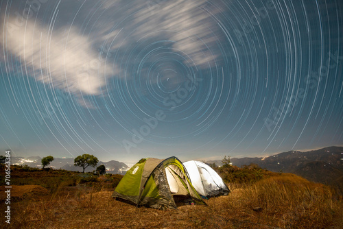 camping in the mountains on two touristic tent in the starry night with the star trail in the sky with bit white clouds with the city backdrop 