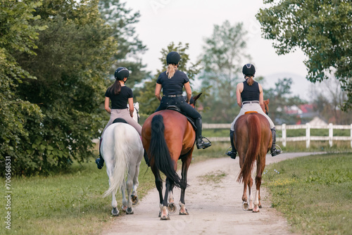 Rear view of three female riders riding horses side by side near white wood fencing, returning to the horse farm
