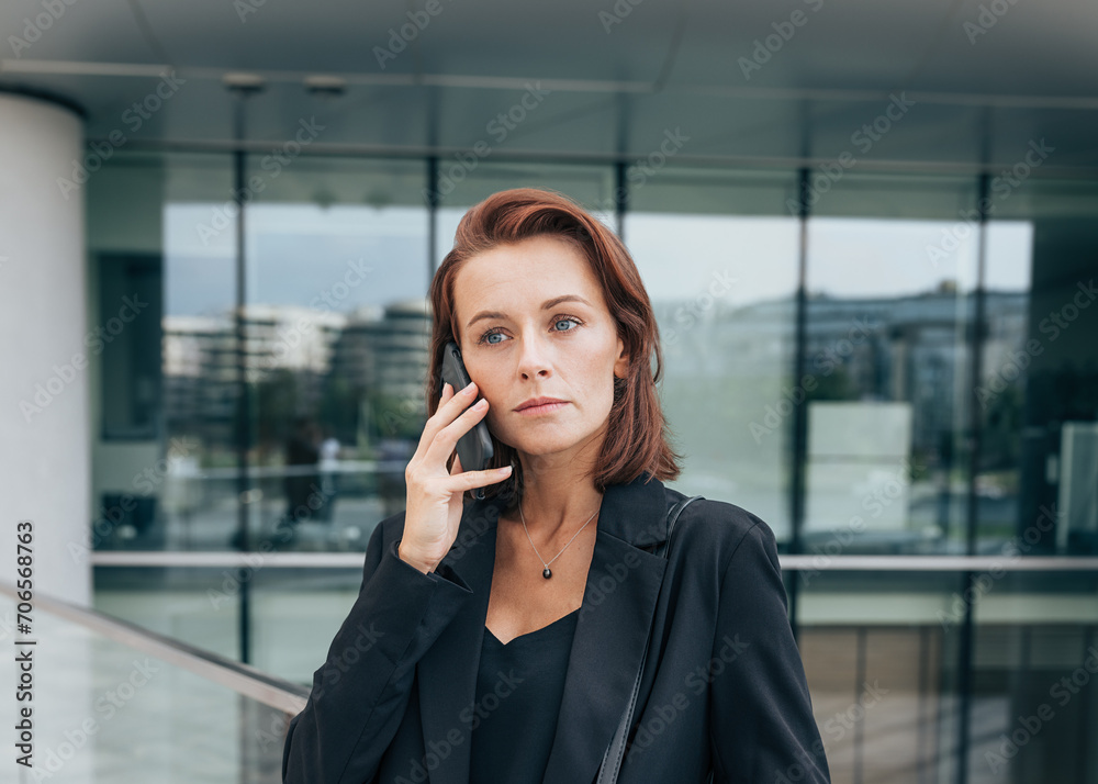 Portrait of a middle-aged female with ginger hair talking on her mobile phone while standing outdoors against a glass building Portrait of a middle-aged female with ginger hair talking on her mobile phone while standing outdoors against a glass building