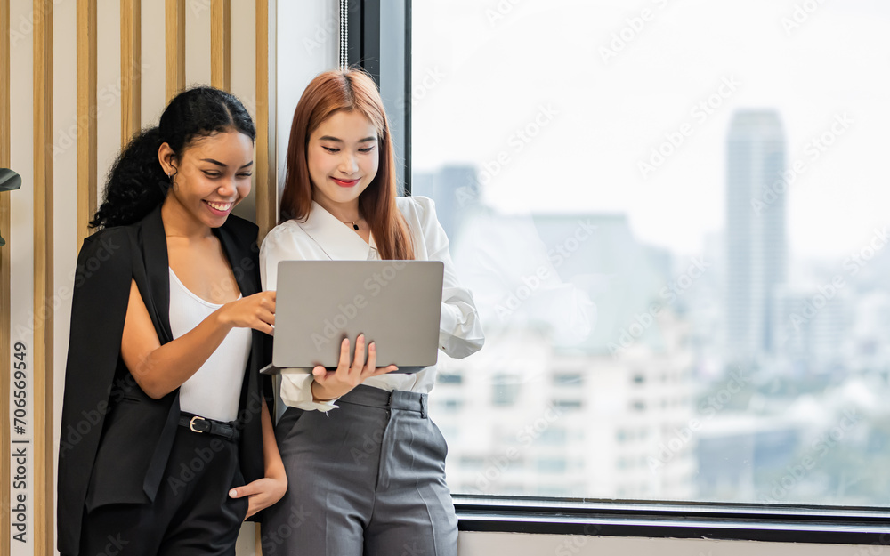 Two diverse mixed race beautiful african and asian business women wearing casual clothes, smiling with happiness, talking while taking break in indoor office, looking at laptop. Two diverse mixed race beautiful african and asian business women wearing casual clothes, smiling with happiness, talking while taking break in indoor office, looking at laptop.