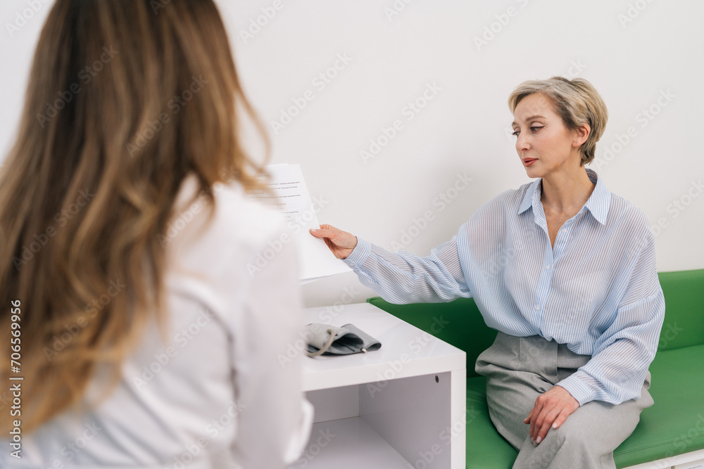 Rear view of unrecognizable woman doctor in white uniform giving paper prescriptions to stressed middle-aged female patient, talking during personal consultation at clinic. Concept of healthcare. Rear view of unrecognizable woman doctor in white uniform giving paper prescriptions to stressed middle-aged female patient, talking during personal consultation at clinic. Concept of healthcare.