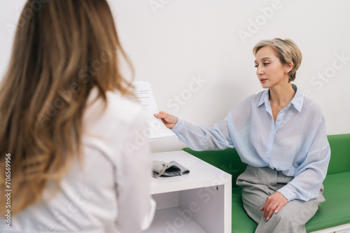 Rear view of unrecognizable woman doctor in white uniform giving paper prescriptions to stressed middle-aged female patient, talking during personal consultation at clinic. Concept of healthcare.