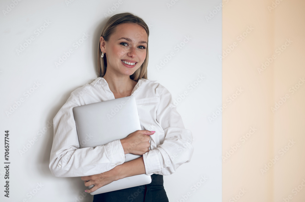 Smiling cute young girl with laptop in hands standing near the wall