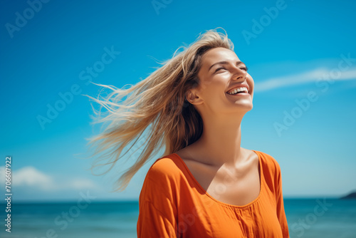 Beautiful young caucasian woman with long blonde hair looking up at the sky smiling, dressed in orange shirt, turquoise water beach background
