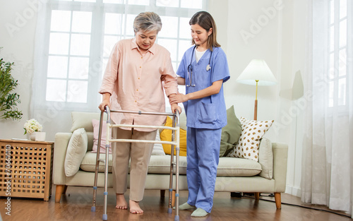 Asian beautiful female nurse or doctor wearing uniform, helping 60s elderly woman to walk by walker, assisting, smiling with happiness, standing in nursing home. Retirement, Healthcare Concept.