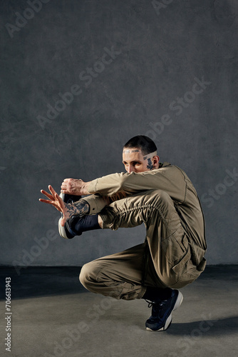 Unusual fellow with tattooed body and face, earrings, beard. Dressed in khaki jumpsuit, black sneakers. Dancing on gray background. Dancehall, hip-hop