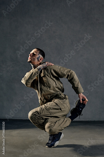 Extraordinary guy with tattooed body, earrings, beard. Dressed in khaki overalls and black sneakers. Dancing on gray background. Dancehall, hip-hop