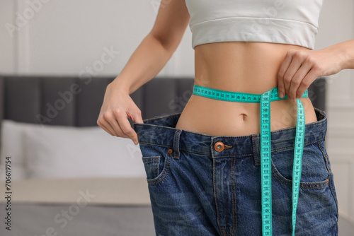 Closeup view of slim woman wearing big jeans and measuring waist with tape in room, space for text. Weight loss