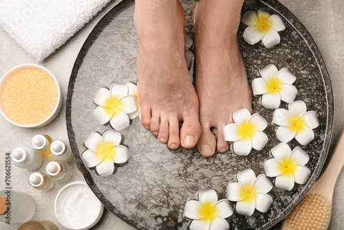 Woman soaking her feet in bowl with water and flowers on light grey floor, top view. Spa treatment