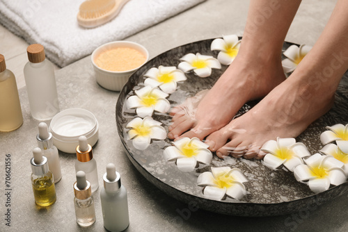Woman soaking her feet in bowl with water and flowers on light grey floor, closeup. Spa treatment