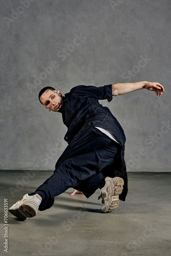Flexible male, tattooed body, beard. Dressed in white t-shirt and sneakers, black denim shirt, pants. Dancing on gray background. Dancehall, hip-hop