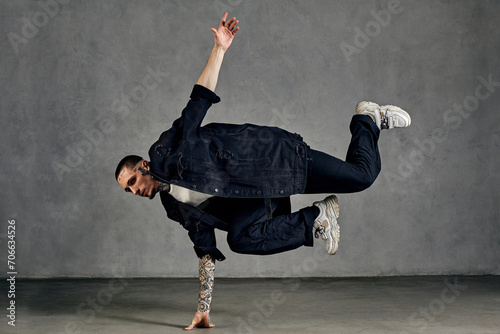 Active male, tattooed body, beard. Dressed in white t-shirt and sneakers, black denim shirt, pants. Dancing on gray background. Dancehall, hip-hop