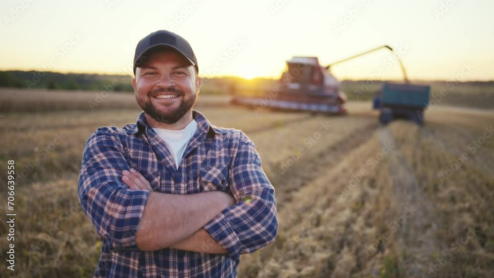 Happy bearded farmer in cap looking at camera standing on agricultural wheat field with combine harvester and dump truck on background. Harvesting, farming at sunset. Food production, agribusiness. Happy bearded farmer in cap looking at camera standing on agricultural wheat field with combine harvester and dump truck on background. Harvesting, farming at sunset. Food production, agribusiness.