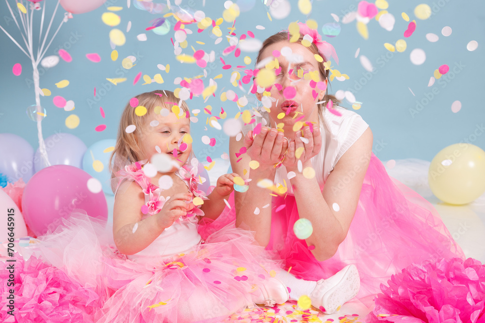 A confetti birthday party celebration. A mother and a little daughter are sitting in pink skirts on blue background.