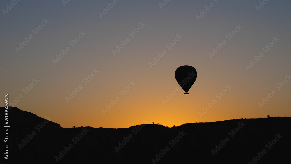 Silhouettes of a hot air balloon hovering over a mountain range. Against the backdrop of the dawn sky. The sun is about to emerge from behind the mountain. Cinematographic shot of a balloon flight.