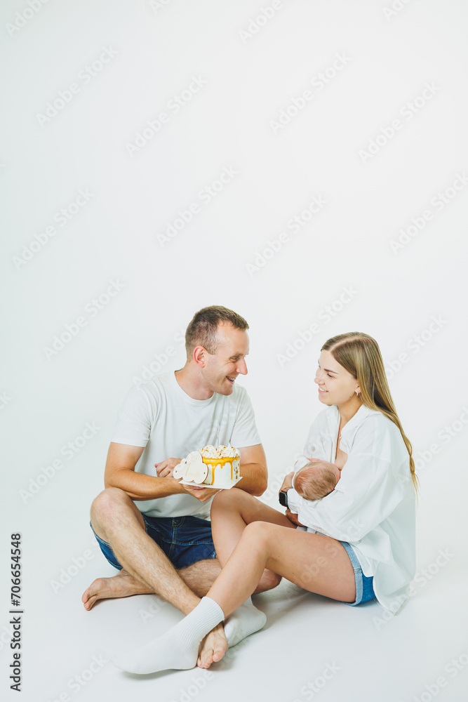 Young happy parents with a baby in their arms and a cake celebrate the first month of the baby's life. Mom and dad with a newborn baby on a white background Young happy parents with a baby in their arms and a cake celebrate the first month of the baby's life. Mom and dad with a newborn baby on a white background