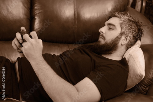 Happy guy using smartphone, enjoying leisure at home, relaxing on leather couch, overusing mobile phone, texting message, chatting online. Cellphone addict, social media obsess photo
